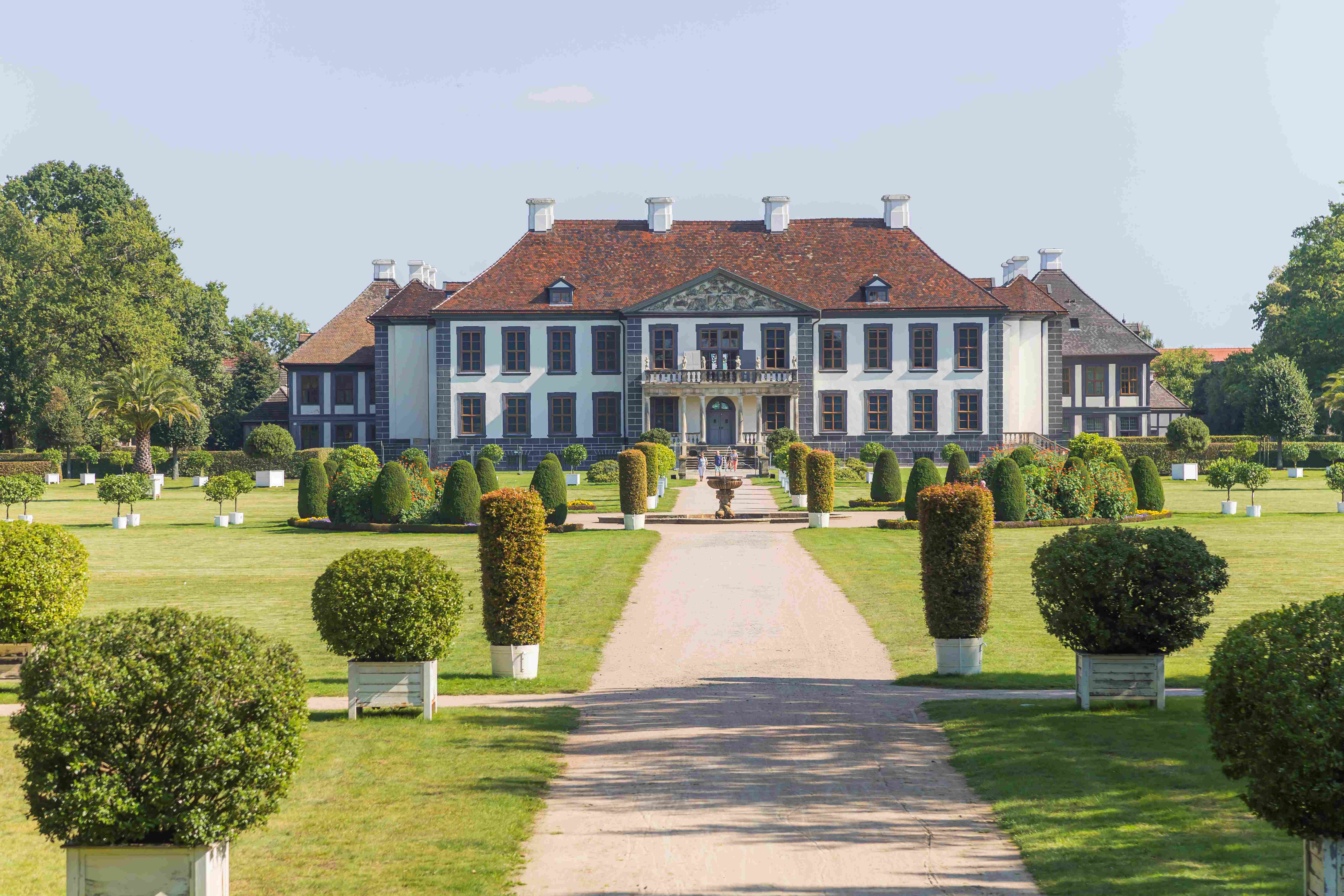 Das Schloss Oranienbaum. Ein zentraler Weg führt direkt auf die schwarz-weiße Fassade des Schlosses zu. An den Seiten stehen Buchsbäume.
