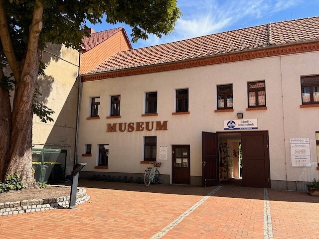 Das Stadtmuseum Oschersleben von Außen. Eine weiße Hausfassade trägt die Aufschrift "Museum". Über einer großen Pforte hängt ein Schild mit dem Verweis zur Stadtbibliothek.