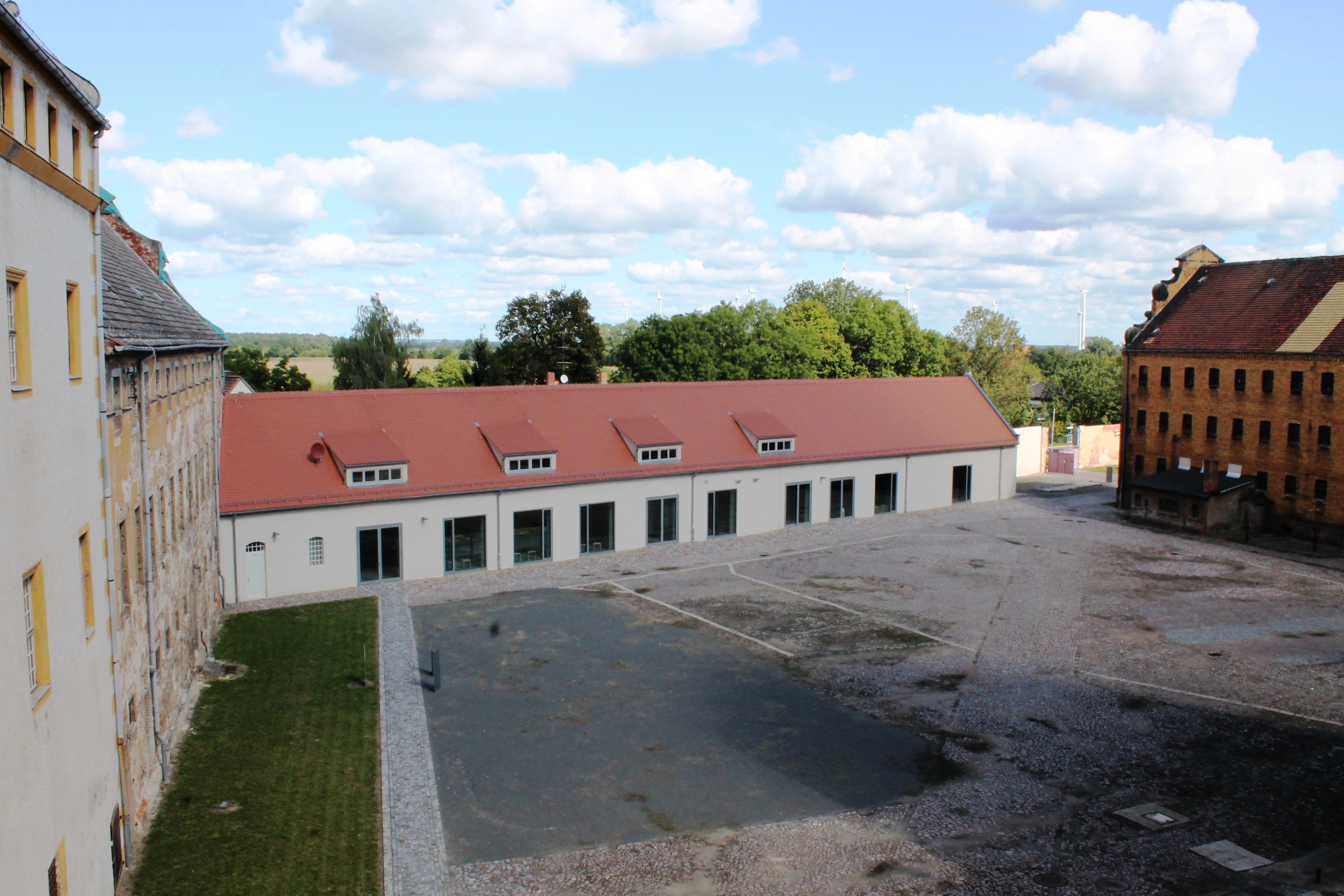 Sammlung Gedenkstätte KZ Lichtenburg Prettin. Um einen grauen Innenhof befinden sich drei Gebäude. Eines davon ist länglich mit einem roten Ziegeldach, eines besteht aus rotem Backstein und das größte ist weiß mit gelben Fensterläden.