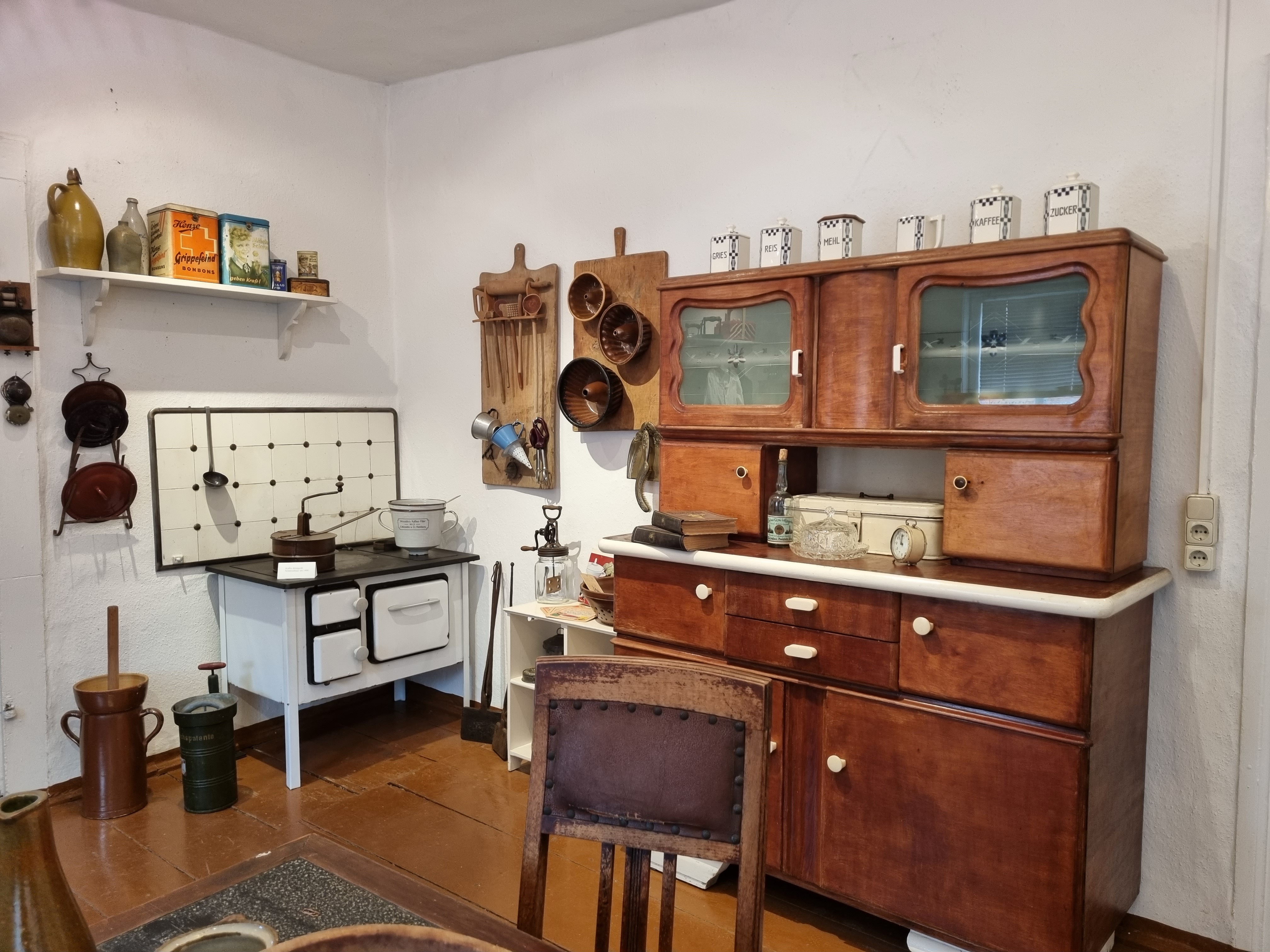 Eine eingerichtete Wohnküche im Heimatmuseum Pretzsch Elbe. Ein weißer Herd, ein Butterstampfer, Kuchenforen und ein Holzbuffet sind Teil der Küche.