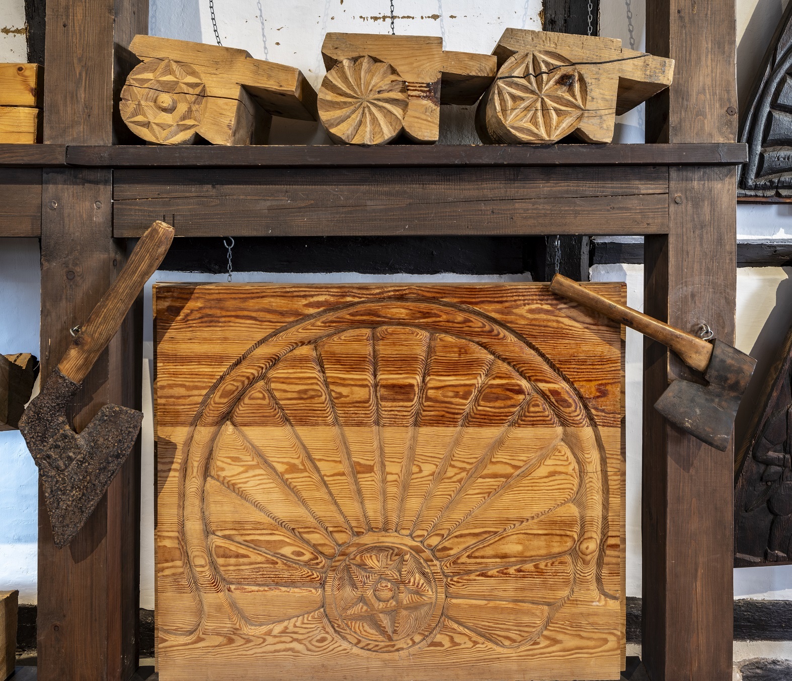 Eine Holzplatte, in die eine Musterung gehauen wurde. An den Seiten hängen Holzbeile und darüber liegen weitere Bauelemente aus Holz, welche Blumen- oder Sternformen besitzen.