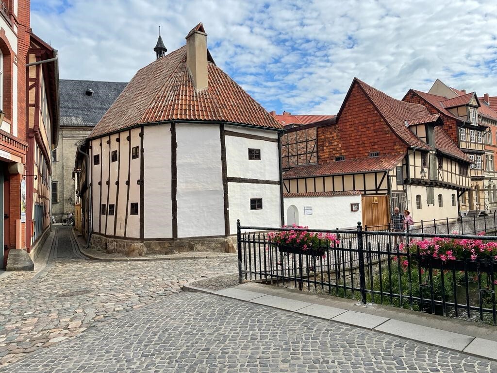 Ständerbau 2022. Der Ständerbau besitzt eine weiße Fachwerkfassade mit einem roten Ziegeldach. Die Fassade wird an der Straßenecke rund weitergeführt. Entlang der Straße sind weitere Fachwerkhäuser erkennbar.