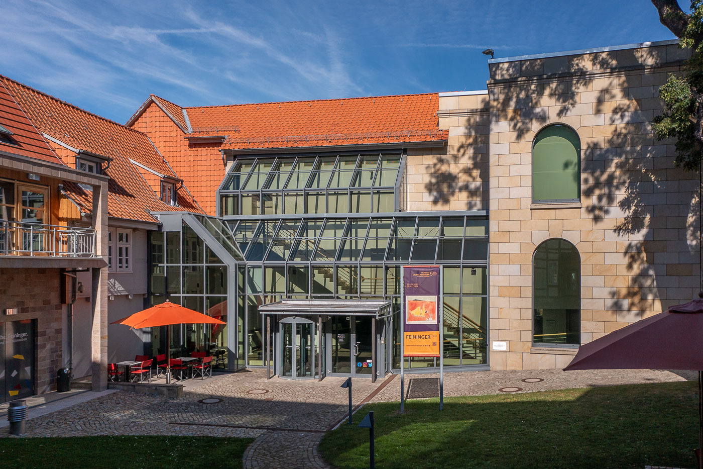 Die Fassade des Lyonel Feininger Museums in Quedlinburg. Der Eingangsbereich besitzt einen Glasbau. Im Innenhof befindet sich ein orangener Schirm mit roten Stühlen darunter.