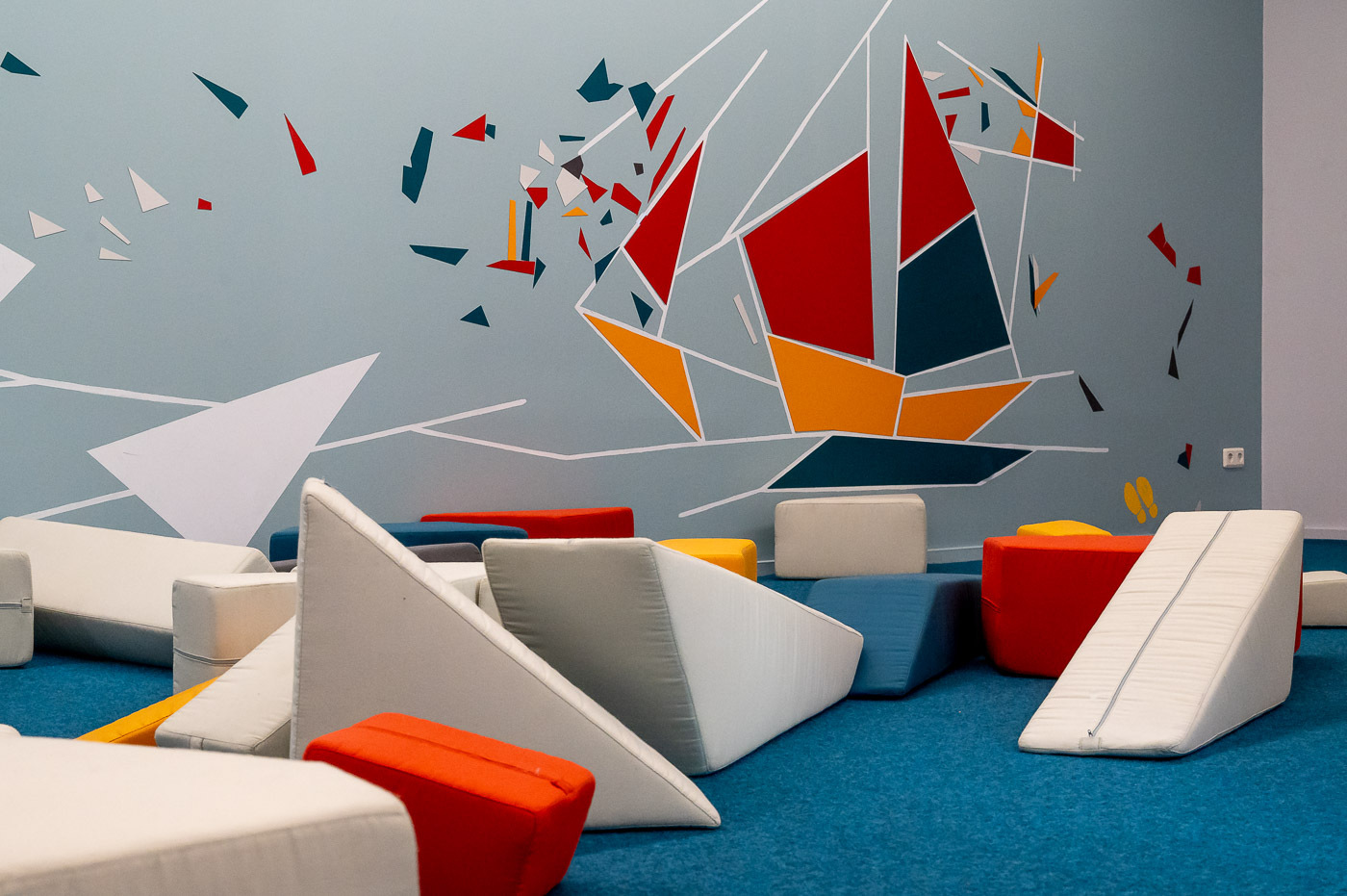 Blick in die Ausstellung "Form, Farbe, Feininger". Auf einem blauen Teppichboden liegen weiße, blaue, rote und gelbe Sitzmöbel in unterschiedlichen Formen, wie Dreiecke und Quader.