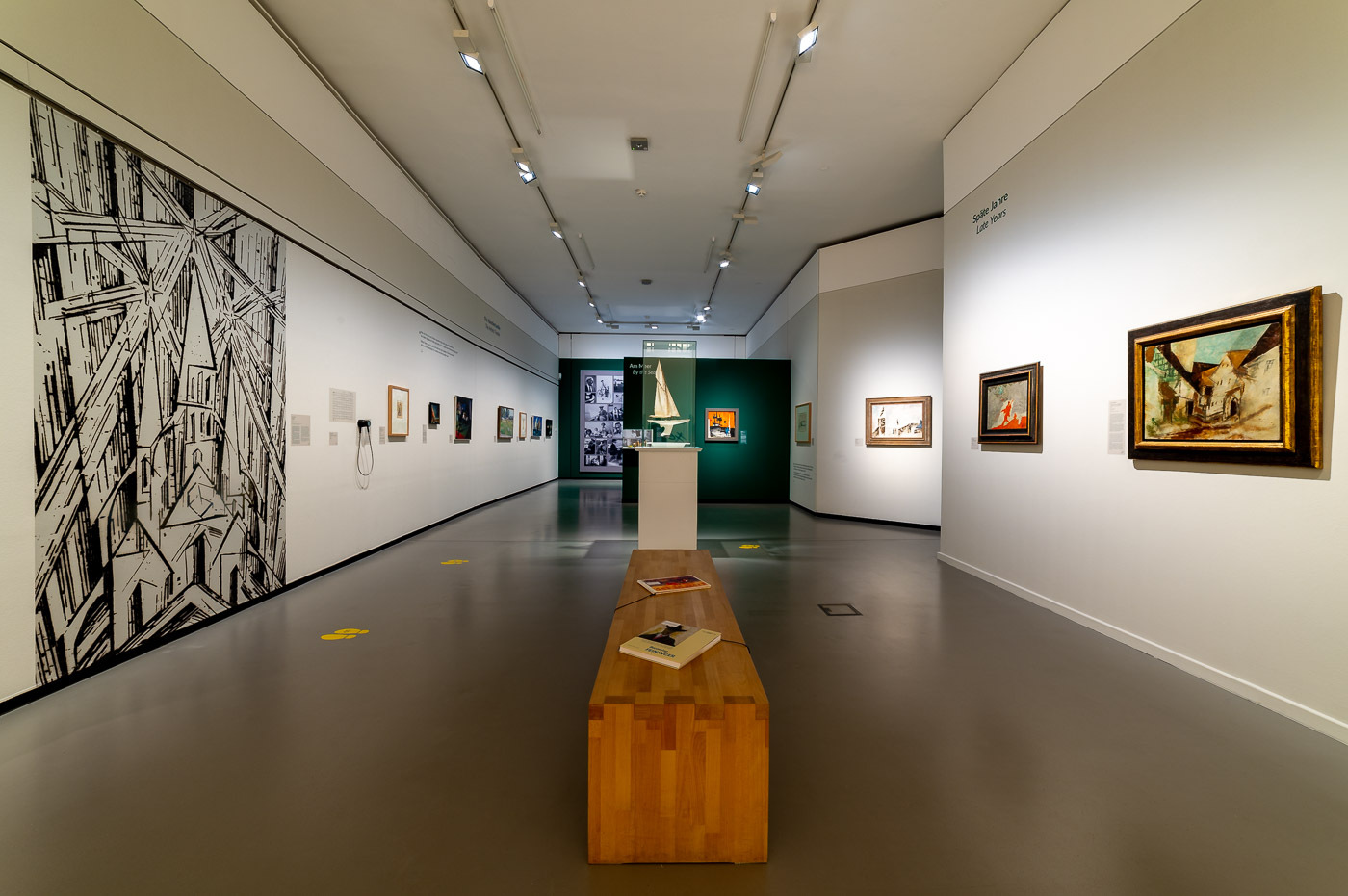 Blick in die Ausstellung "Lyonel Feininger. Meister der Moderne". In der Mitte des Raumes steht eine Holzbank. An den weißen Wänden zu beide Seiten hängen Feiningers Gemälde in unterschiedlichen Formaten. Auf der linken Seite nimmt eine schwarz-weiße Grafik die gesamte Höhe der Wand ein.