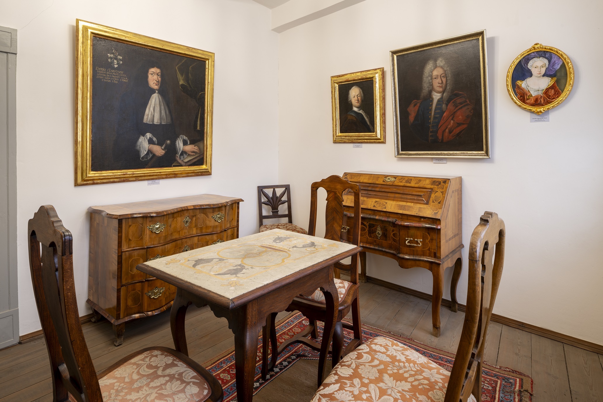 Ein Ausstellungraum mit Holzinterieur. Ein schmaler hoher Tisch in der Mitte des Raumes besitzt eine Marmorplatte und steht auf einem roten Teppich. Drei Polsterstühle stehen um den Tisch. An den Wänden stehen weitere Holzmöbel und vier unterschiedlich große Portraitgemälde sind darüber angebracht.