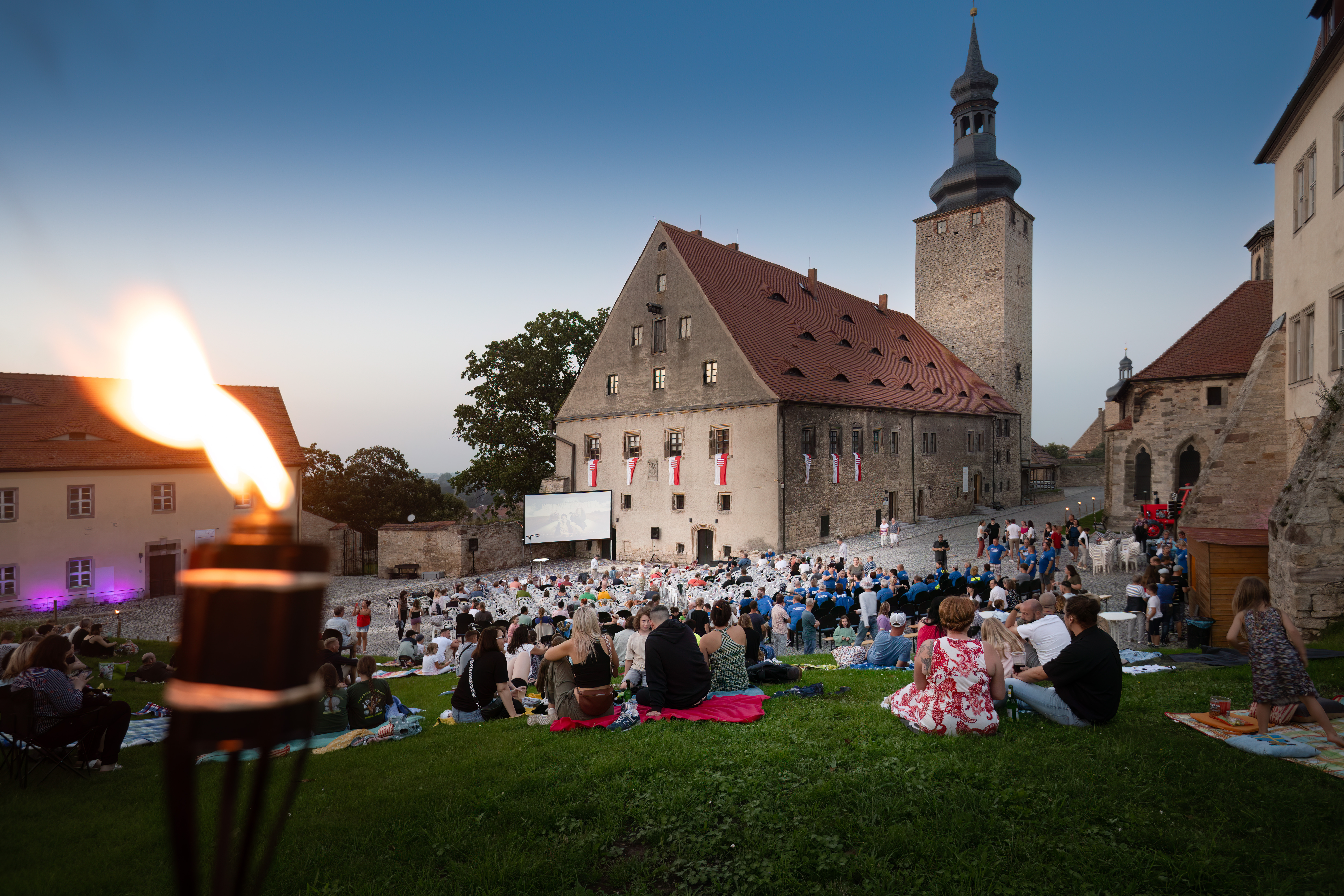 Sommerkino auf der Burg Querfurt. Besucher sitzen auf einer Wiese und schauen auf eine Leinwand, die zwischen den Burggebäuden aufgestellt wurde.