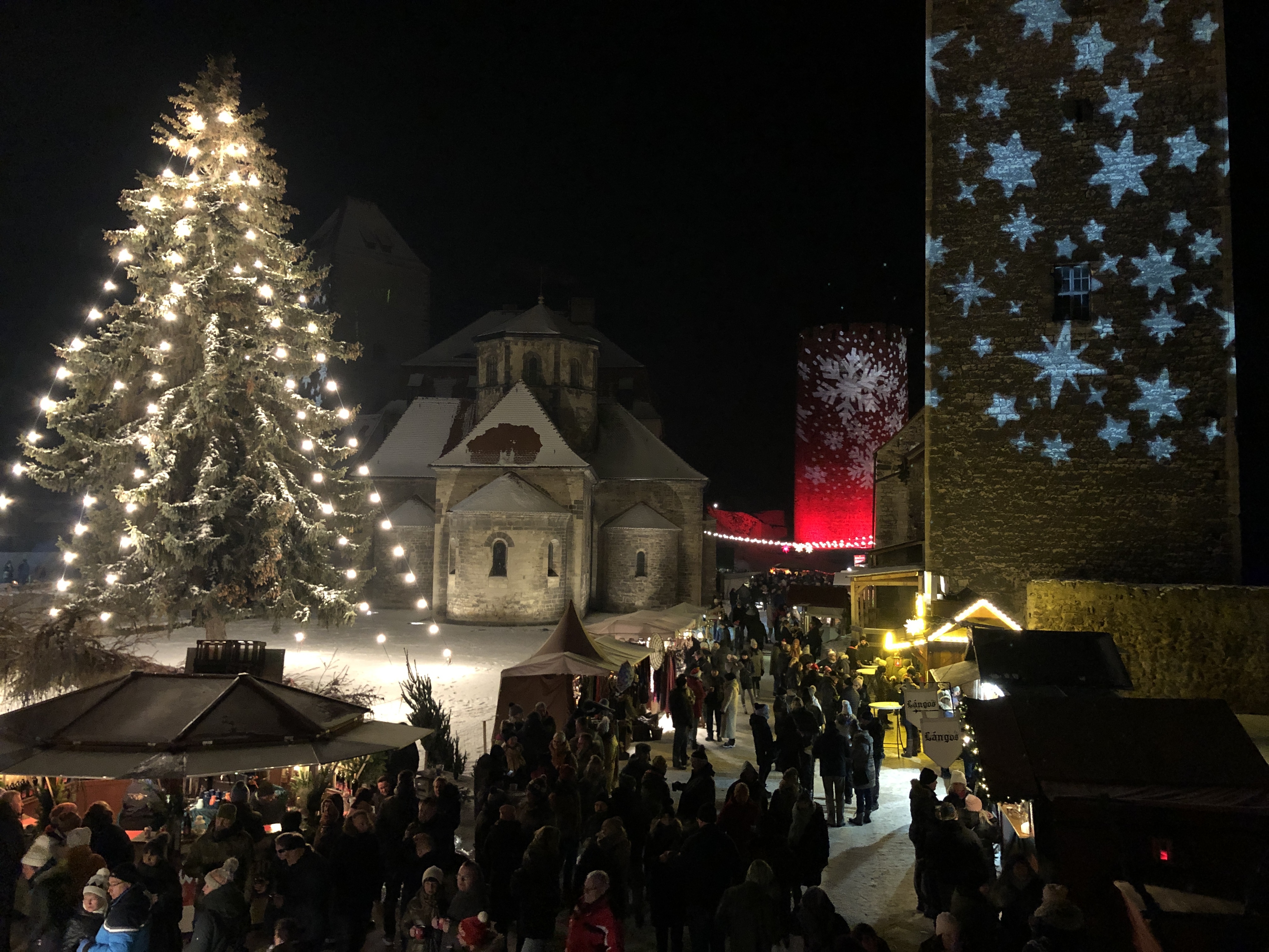 Weihnachtszauber. Weihnachtsmarkt auf der Burg Querfurt. Ein großer mit Lichtern geschmückter Weihnachtsbaum steht in der Mitte des mit Schnee bedeckten Innenhofes. Darum herum befinden sich Weihnachtsmarktbuden. Die Burg und die Türme werden weihnachtlich illuminiert.