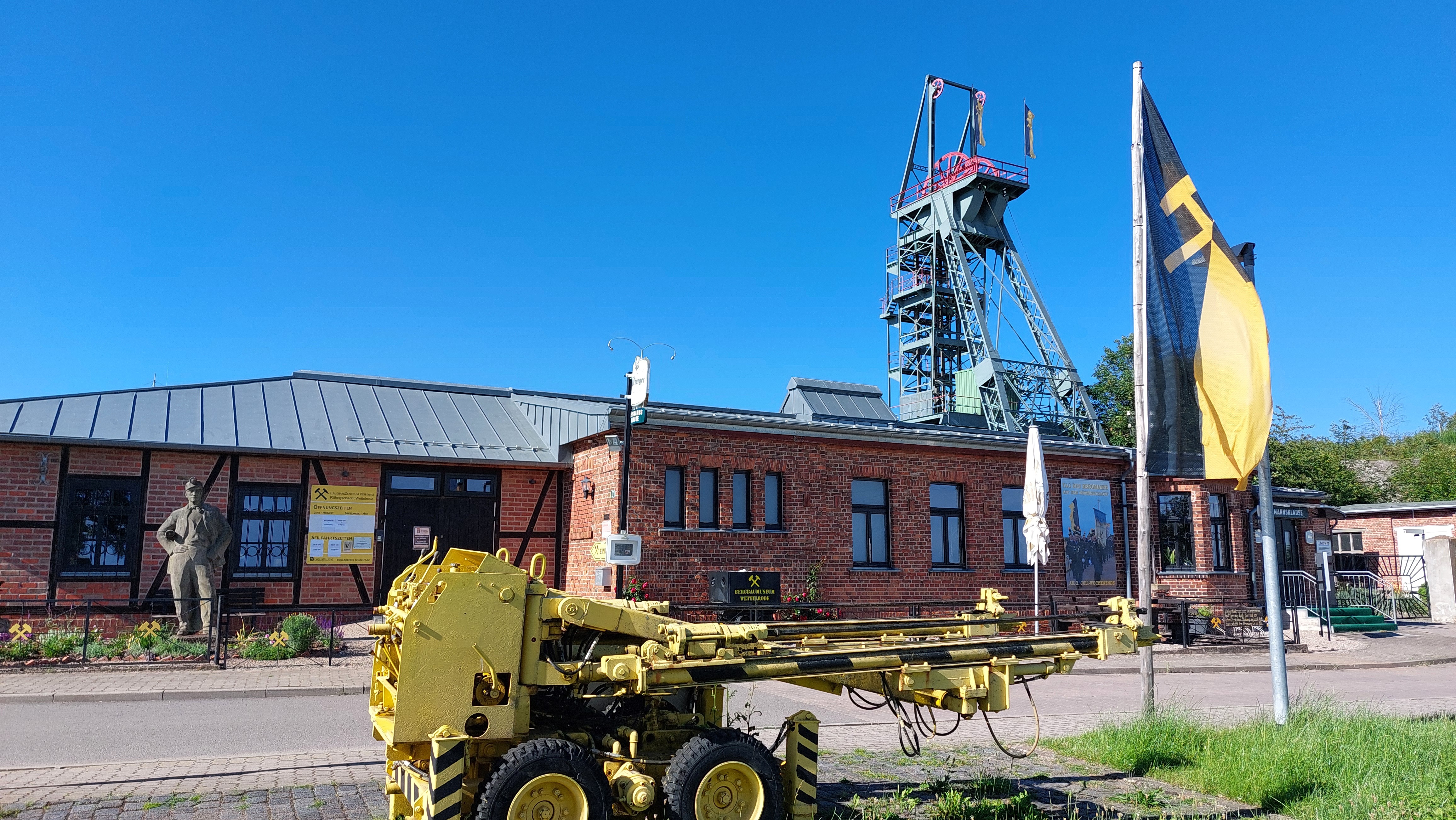 Das ErlebnisZentrum Bergbau Röhrigschacht Wettelrode von Außen. Ein rotes flaches Backsteingebäude, vor dem ein gelbes Gerät für den Bergbau steht.