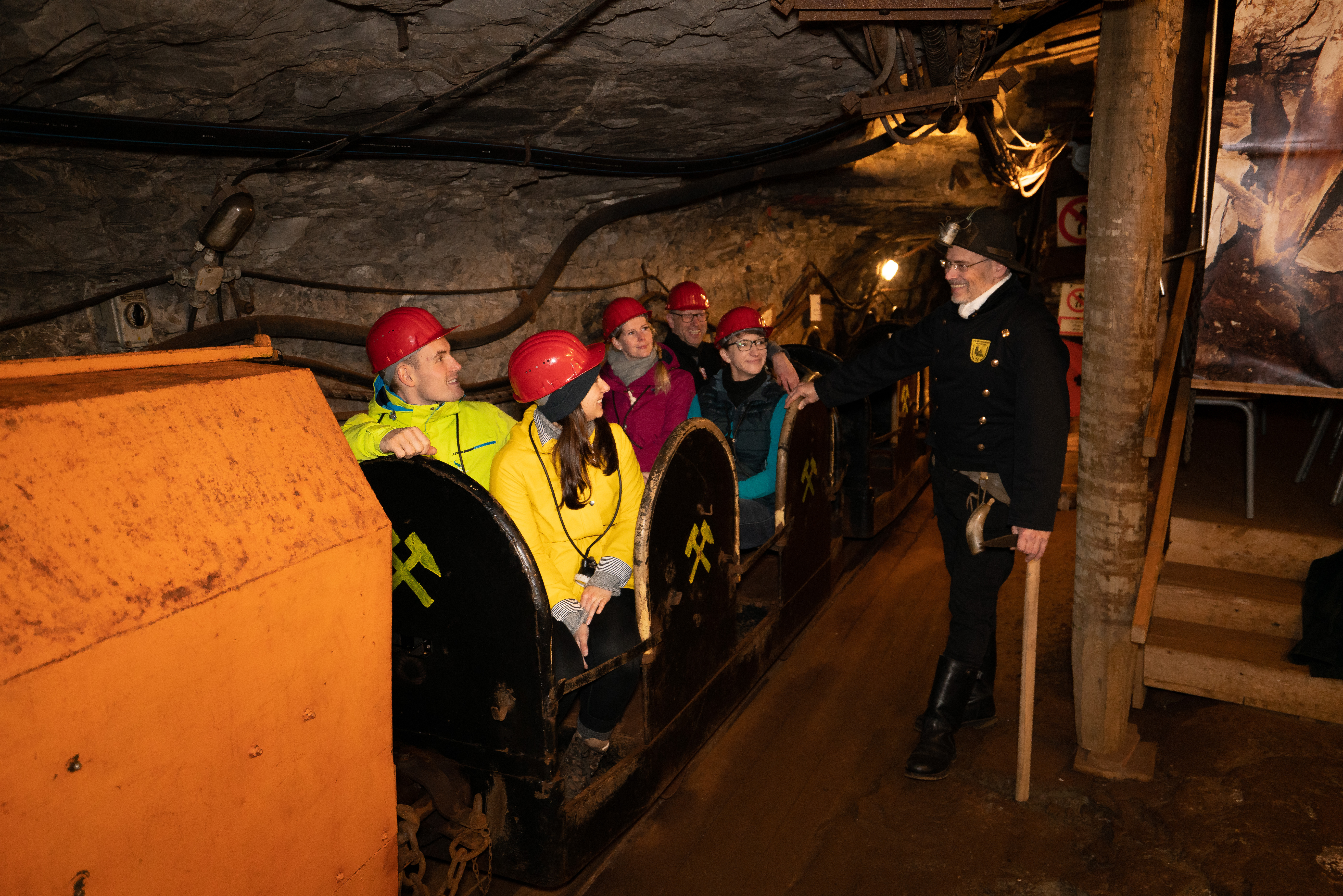 Mehrere Personen sitzen in einer Bahn im Bergwerk. Ein Mitarbeiter in der Uniform eines Bergwerkarbeiters erzählt etwas.
