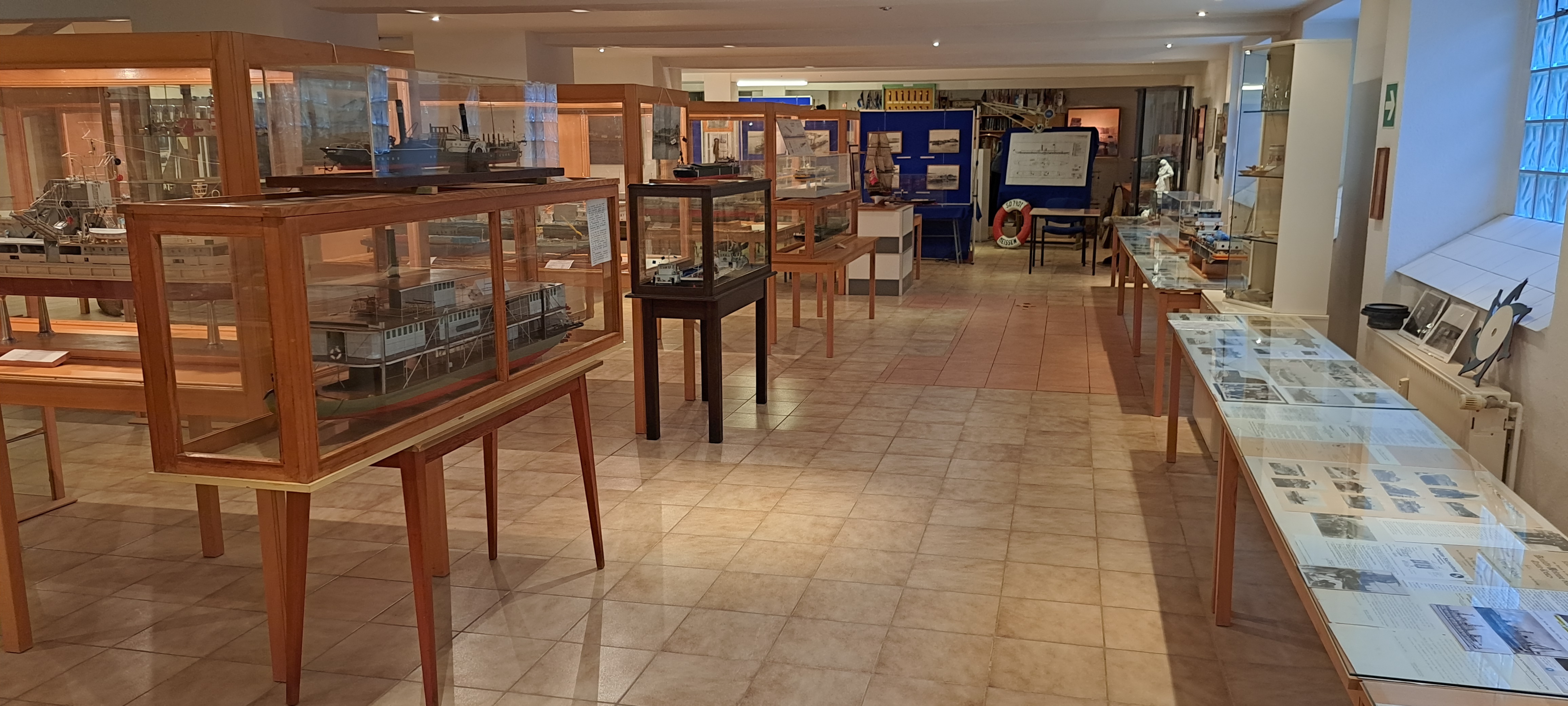 Blick in die Ausstellung: kniehohe Holzvitrinen mit Schiffsmodellen und Informationsmaterialien mit Fotografien befinden sich im Ausstellungsraum.