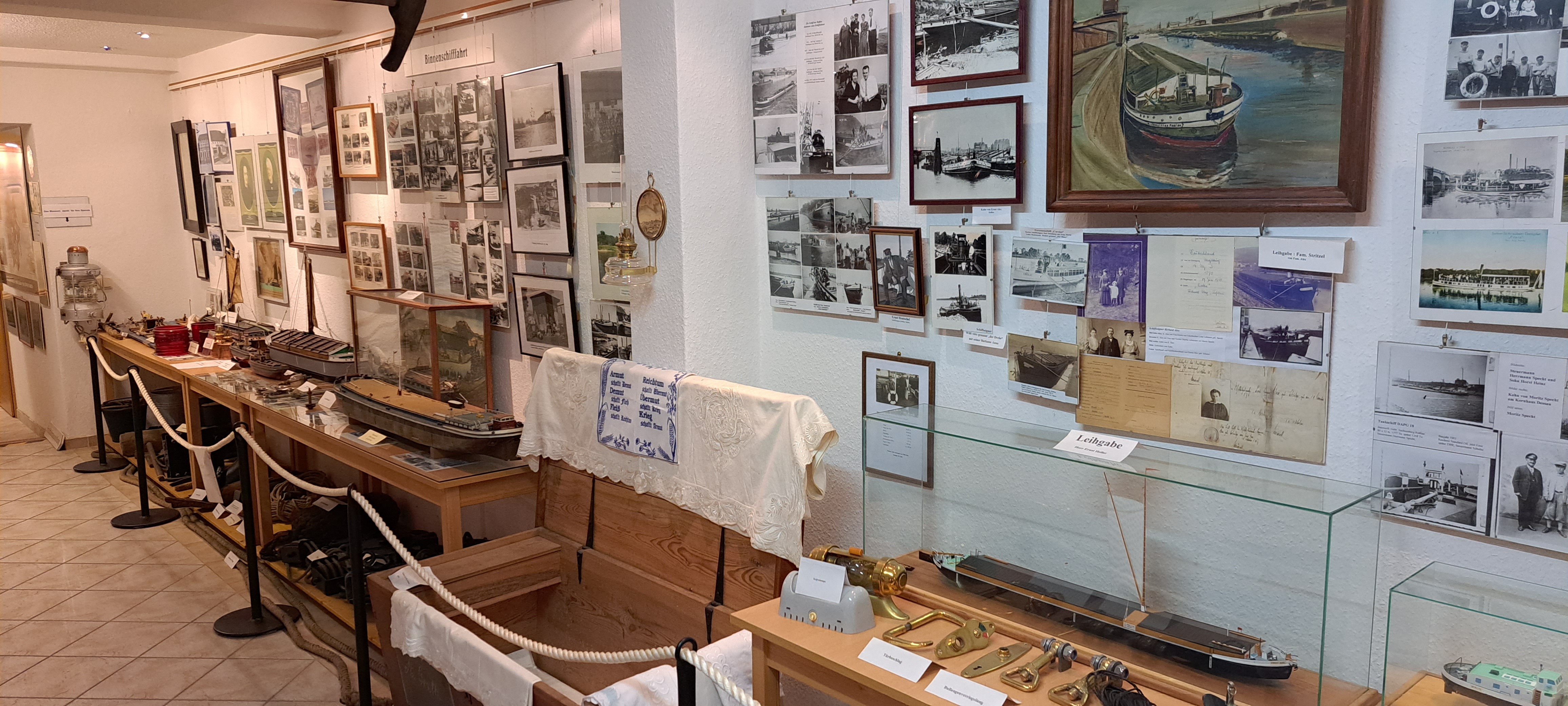 Blick in die Ausstellung: An der Wand hängen verschiedene schwarz-weiß Fotografien und Gemälde von Schiffen. In Vitrinen werden Schiffsmodelle und -zubehör in Regalen darunter ausgestellt