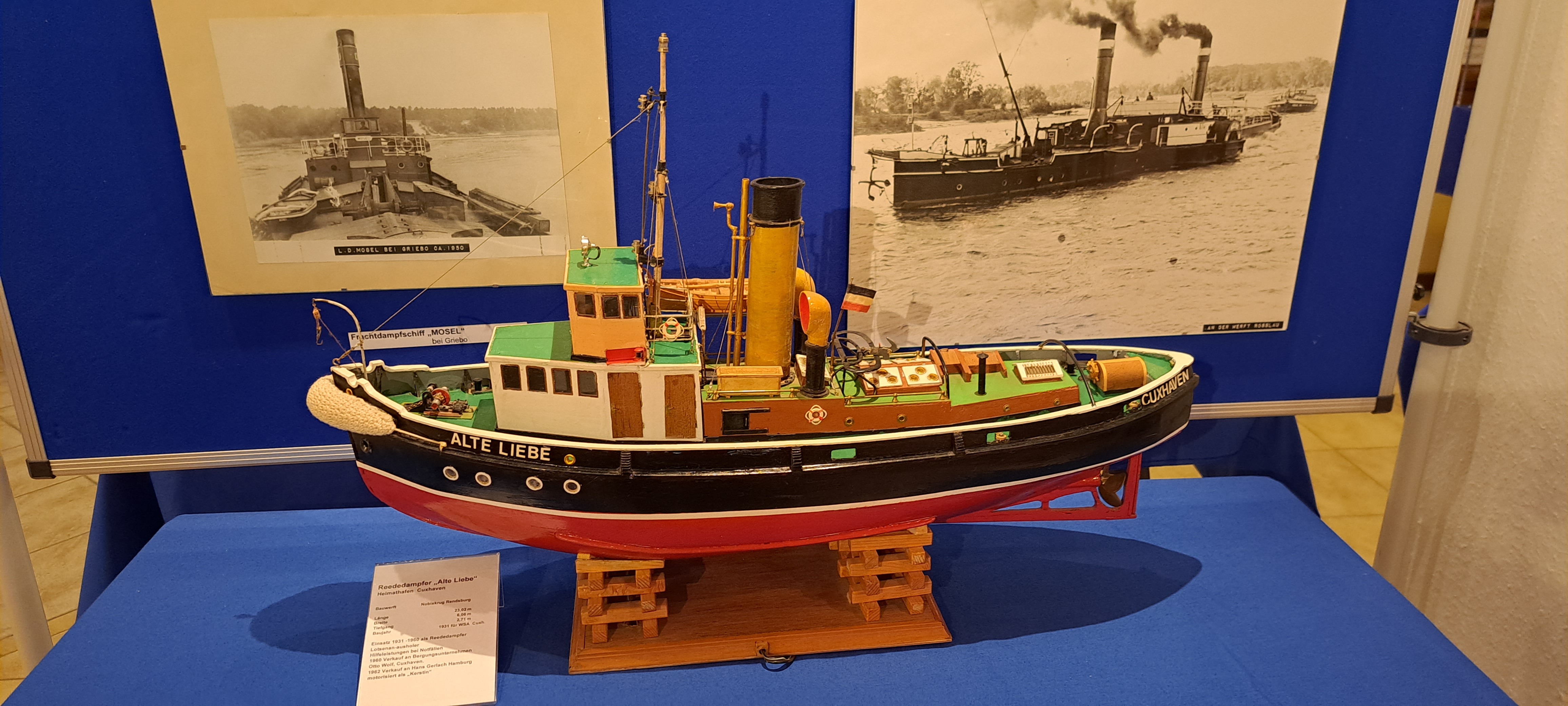 Ein buntes Schiffsmodell mit dem Namen "Ate Liebe". Im Hintergrund sind Fotografien des lebensgroßen Schiffes zu erkennen.