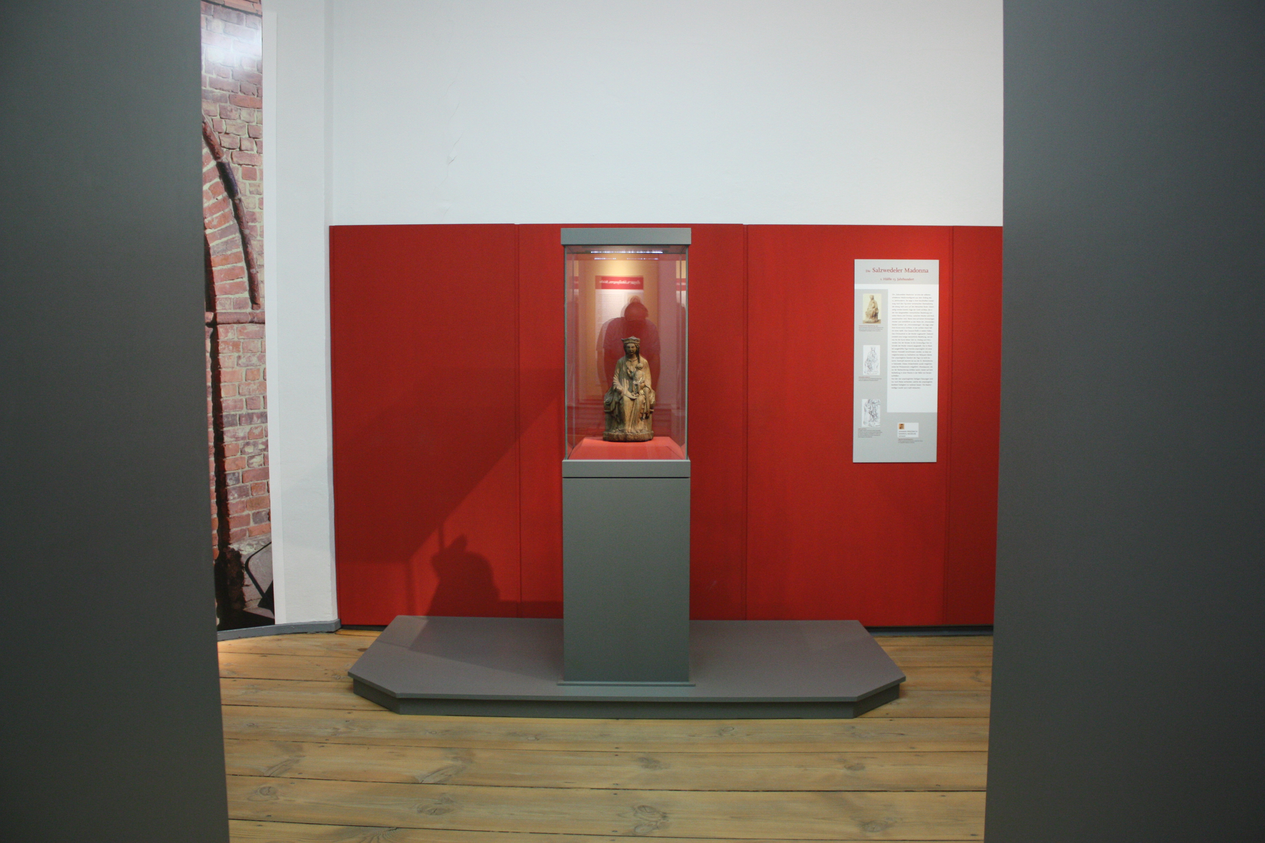 Salzwedeler Madonna. Eine Madonnenstatue aus Holz wird auf einem roten Untergrund in einer Vitrine vor einem roten Hintergrund präsentiert.
