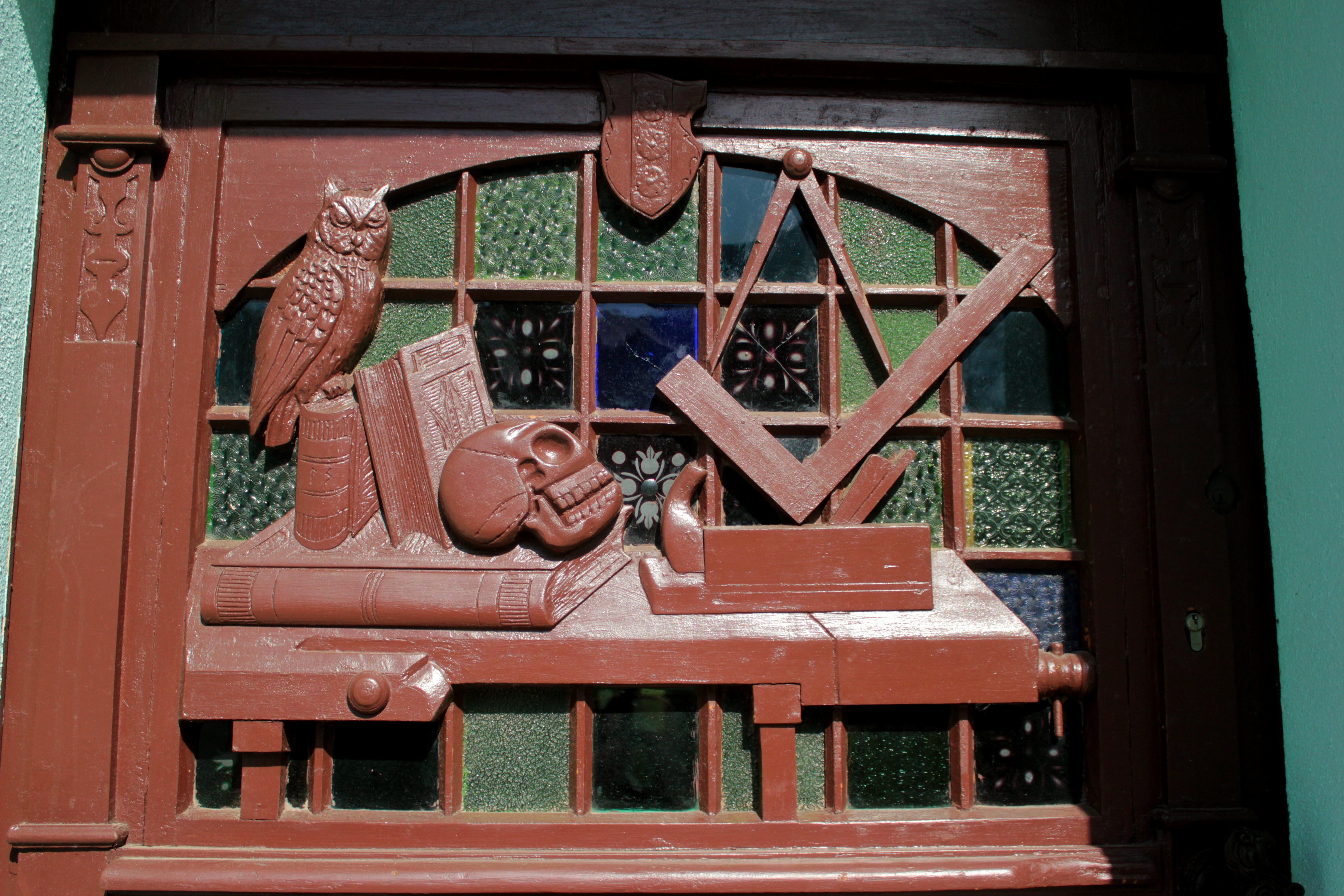 Schnitzerei von Gustav Adolf Spengler an der Haustür des Spengler Hauses. Darauf ist ein Schreibtisch mit mehreren Büchern, einer Eule, einem Totenschädel und einem Zirkel, der über einem Winkel steht.