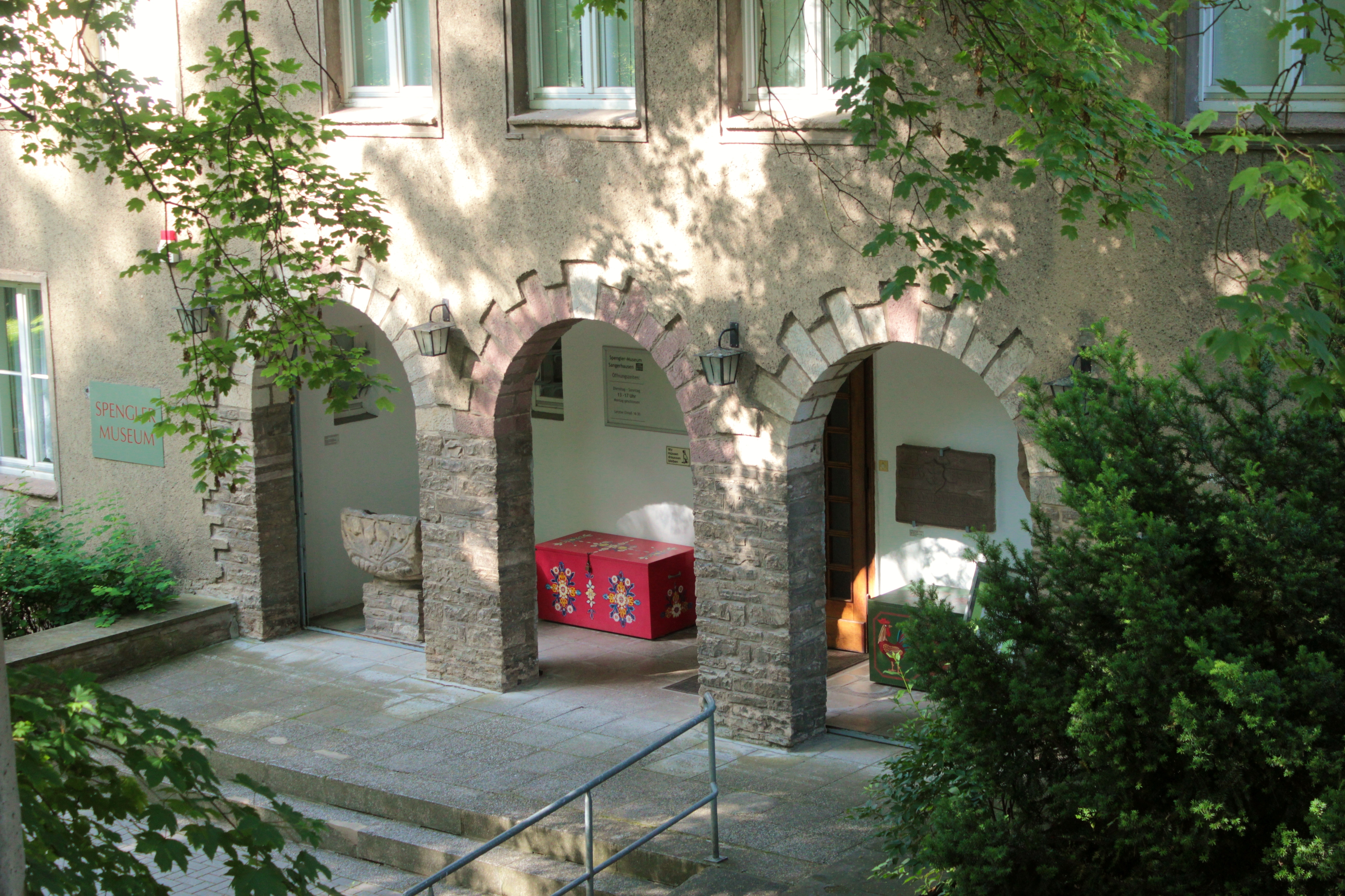 Der Eingangsbereich des 1952 errichteten Museumsgebäude. Drei Arkadenbögen aus Stein und zwei bunte Kisten führen zur Eingangstür des Museums.