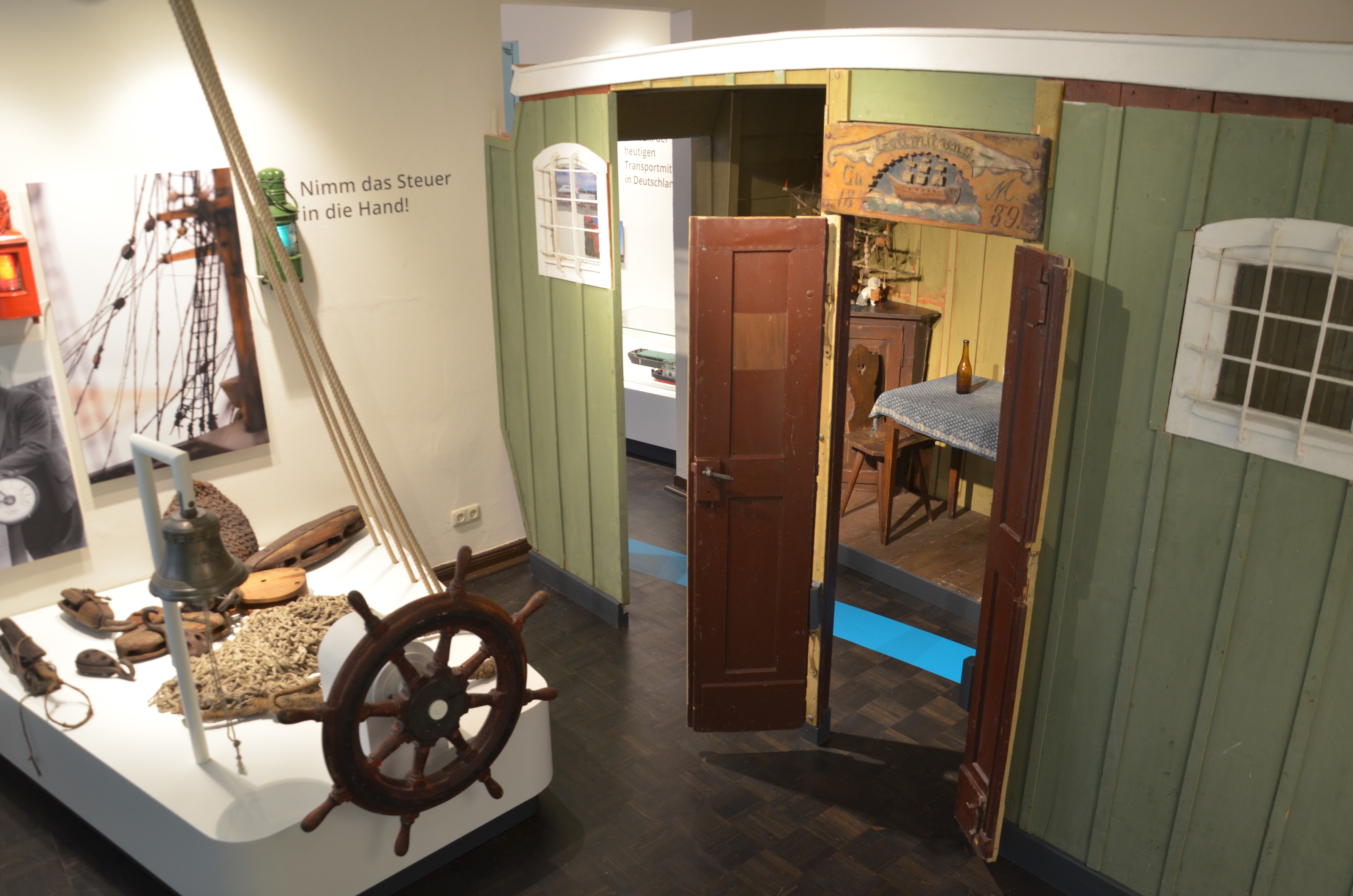 In der Ausstellung "Lebensströme" zeigt den Nachbau einer Bootskajüte, die mit hellgrünem Holz verkleidet ist. Ein dunkles Steuerrad, ein Fischernetz sowie andere Gegenstände werden auf einem weißen Podest präsentiert.