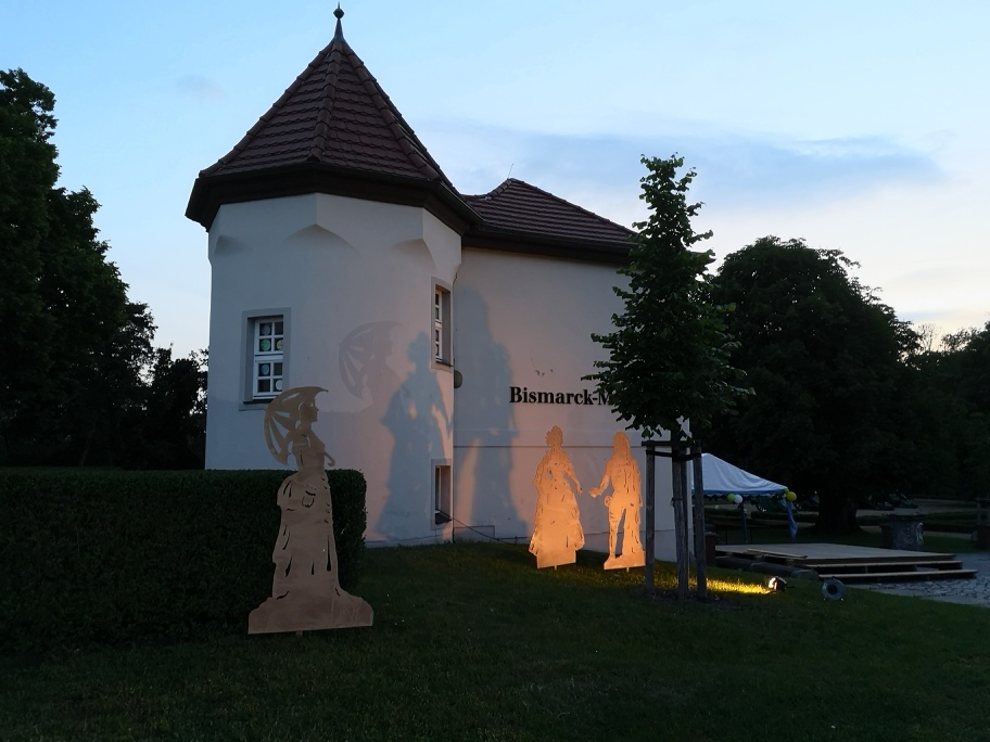 Die historische Gutsanlage in Schönhausen mit dem Seitenflügel des Geburtshauses Otto von Bismarcks. Zwei Figuren stehen vor dem dem Gebäude und werden so beleuchtet, dass ihre Schatten auf die weiße Fassade des Geburtshauses fallen.