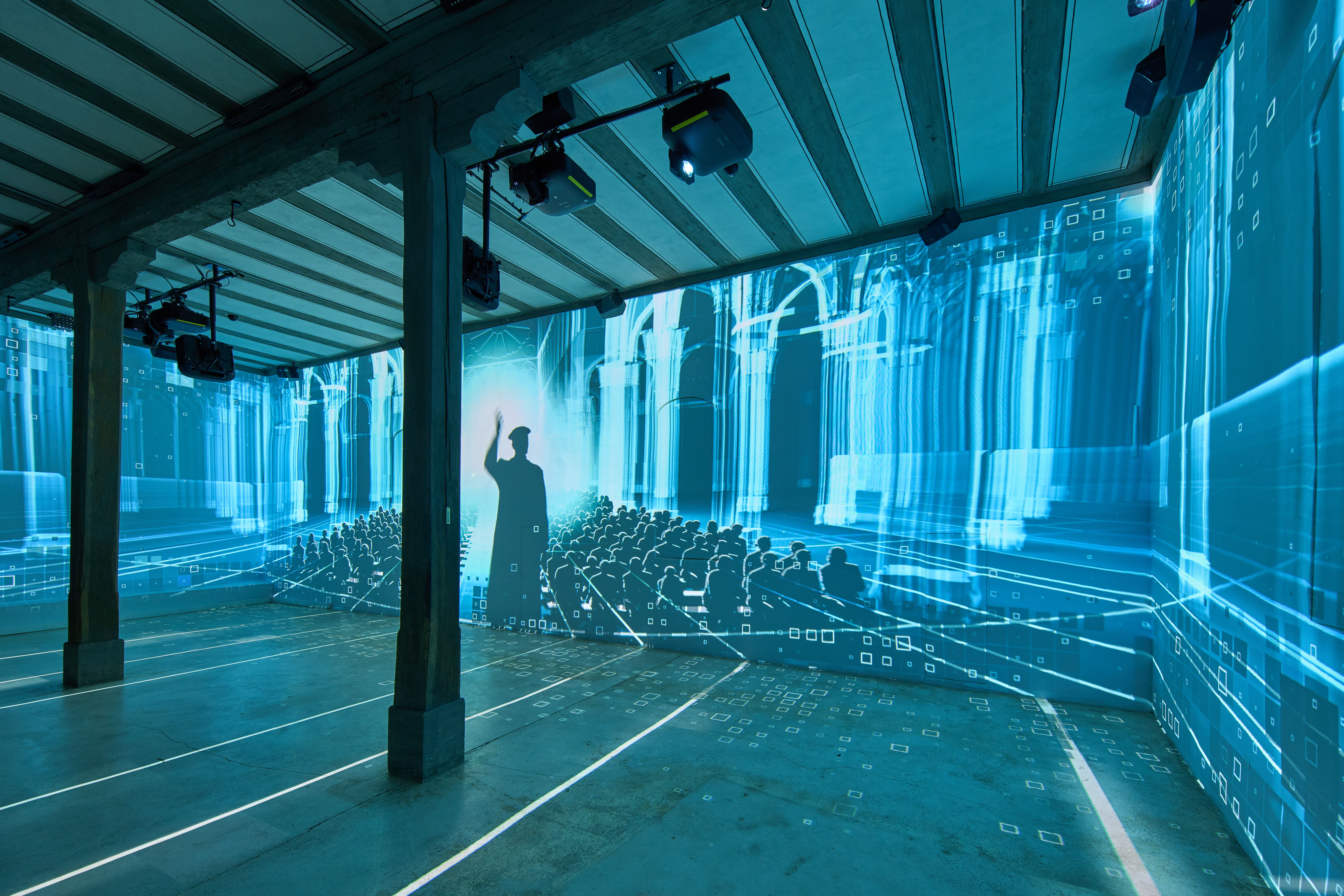 Multimediales Erlebnis in der Hofstube. Auf die Wände eines Raumes wird ein Film projiziert. Die Silhouette eines Mannes mit gehobener Hand vor einer in blaues Licht getauchten kirchlichen Innenarchitektur ist darauf erkennbar.