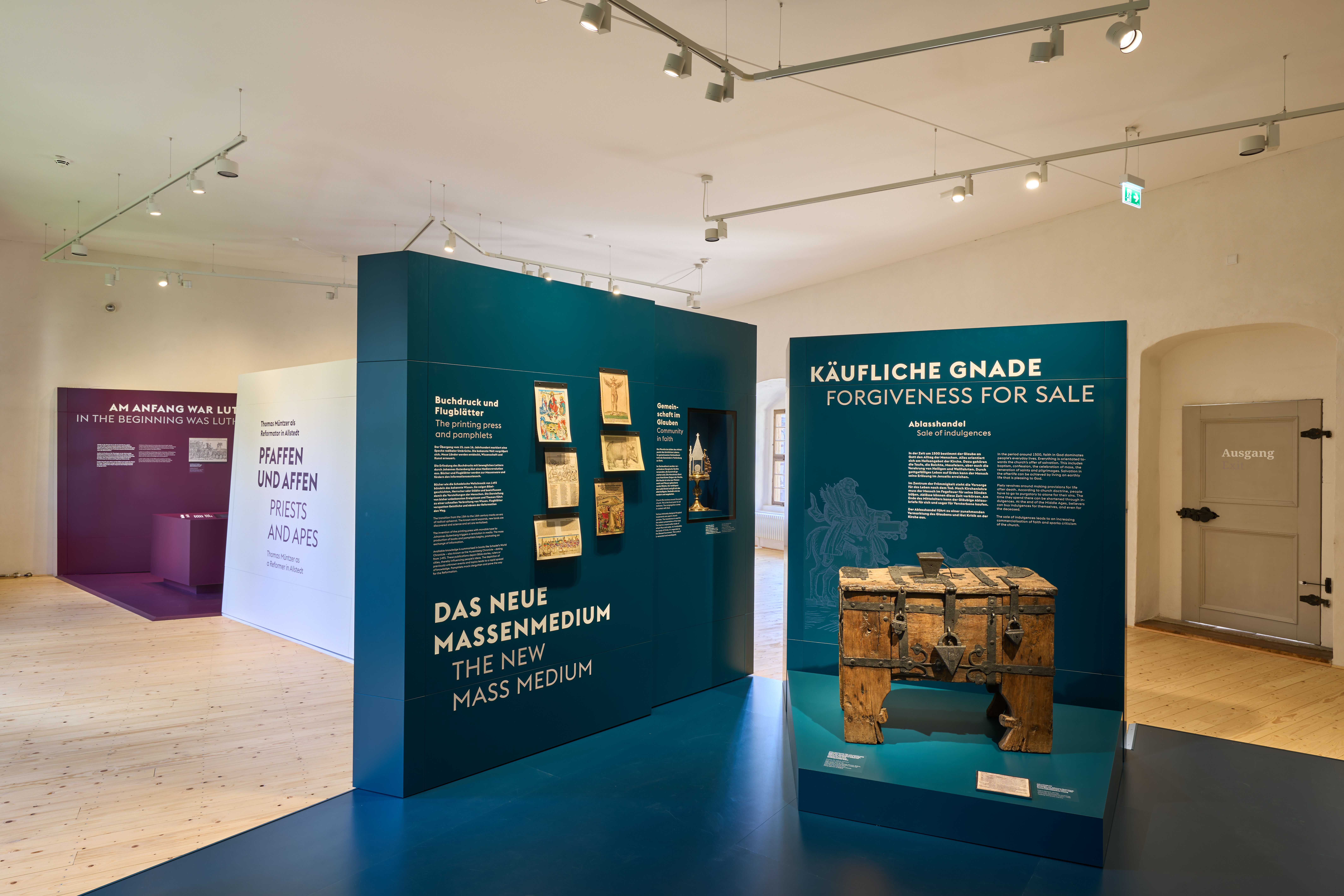 Blick in die Neue Dauerausstellung „Sein und Schein. Thomas Müntzer“. Zwei freistehende blaue Wände präsentieren Texte der Ausstellung. Eine massive Holztruhe steht auf einem Podest vor einer der blauen Wände. Im Hintergrund befinden sich eine weiße und eine violette Wand der Ausstellung.