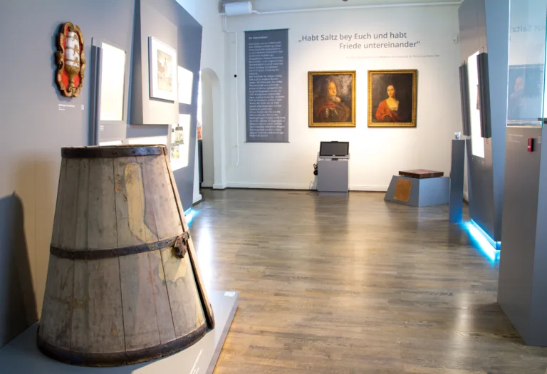 Salzspueren. Ein helles sich nach ober verjüngendes Salzfass steht auf der linken Seite zu Beginn der Ausstellung. Zwei Porträts eines Mannes und einer Frau mit roter Kleidung hängen an der Wand dahinter.