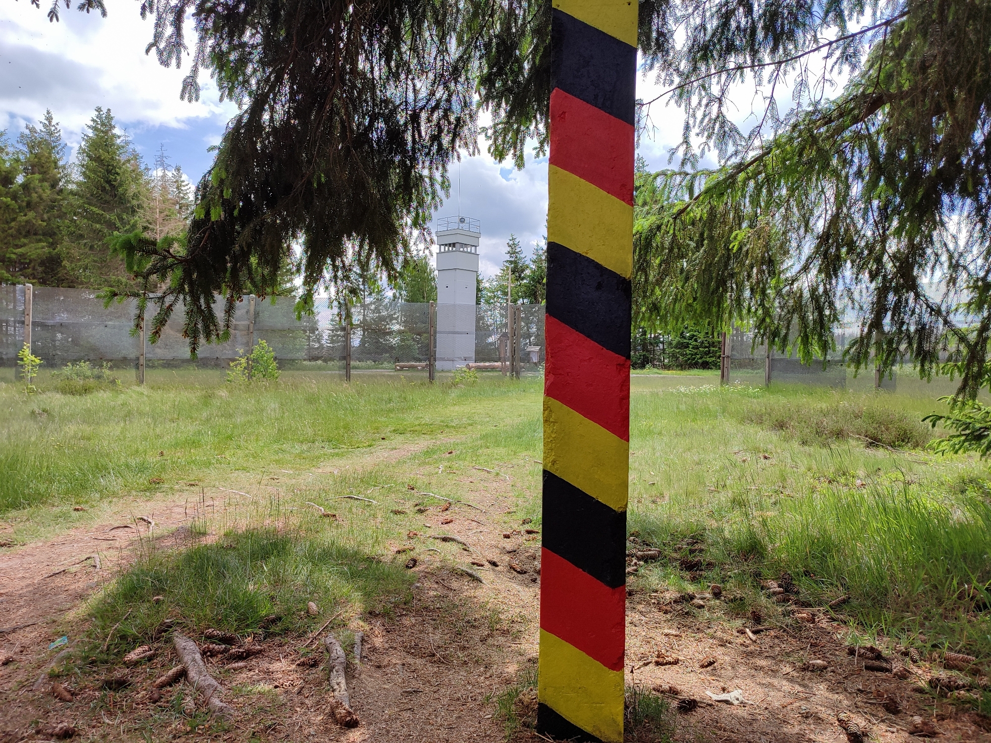 Grenzpfeiler mit den Farben Schwarz, Gelb und Rot. Im Hintergrund wird der Wachturm und Grenzzaun sichtbar.