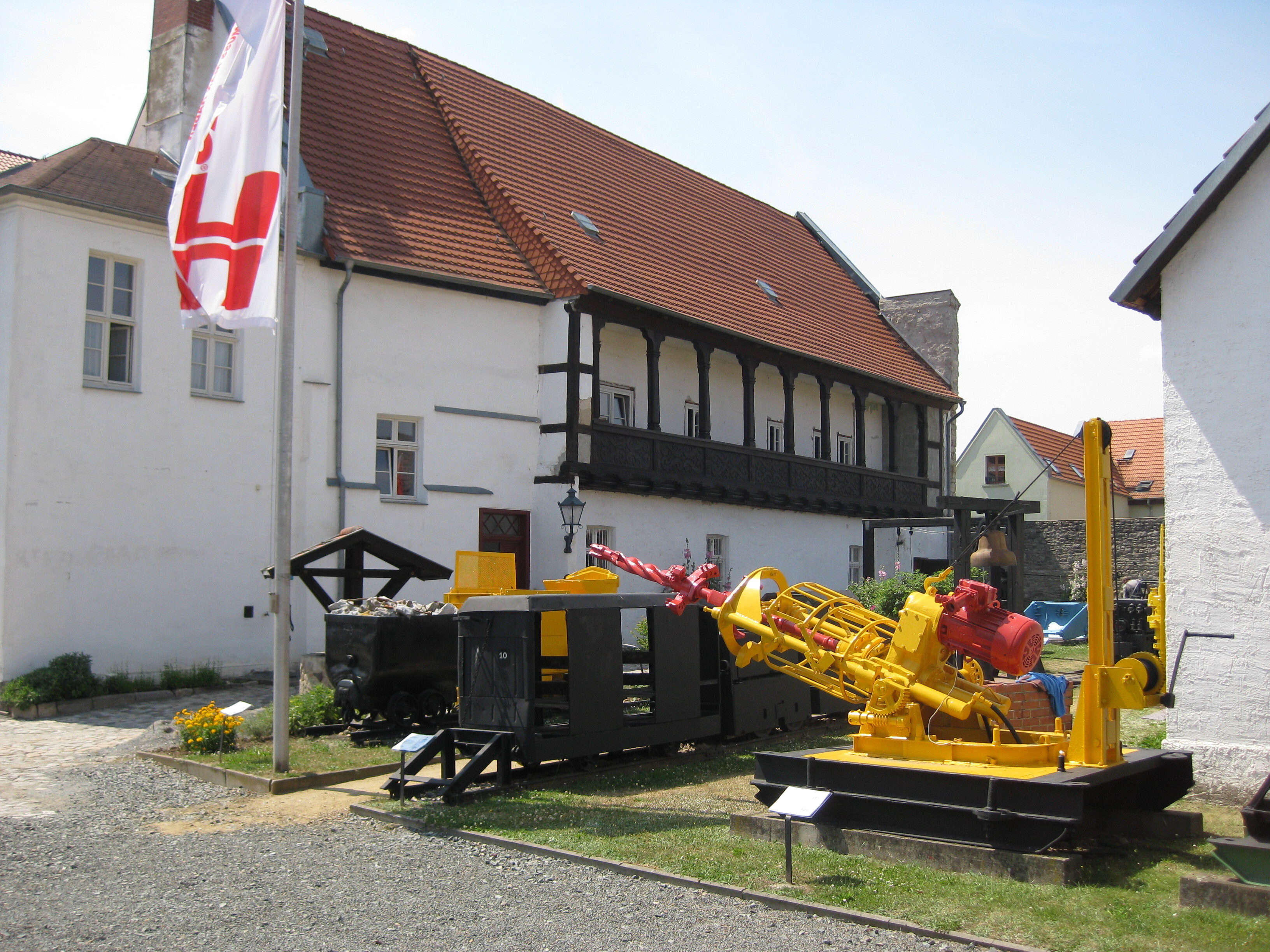 Im Innenhof des Museums steht eine große gelbe Bohrmaschine. Dahinter stehen zwei schwarze Förderwägen, wobei einer mit steinen beladen wurde. Die weiße Fassade des Museums ist dahinter zu sehen.