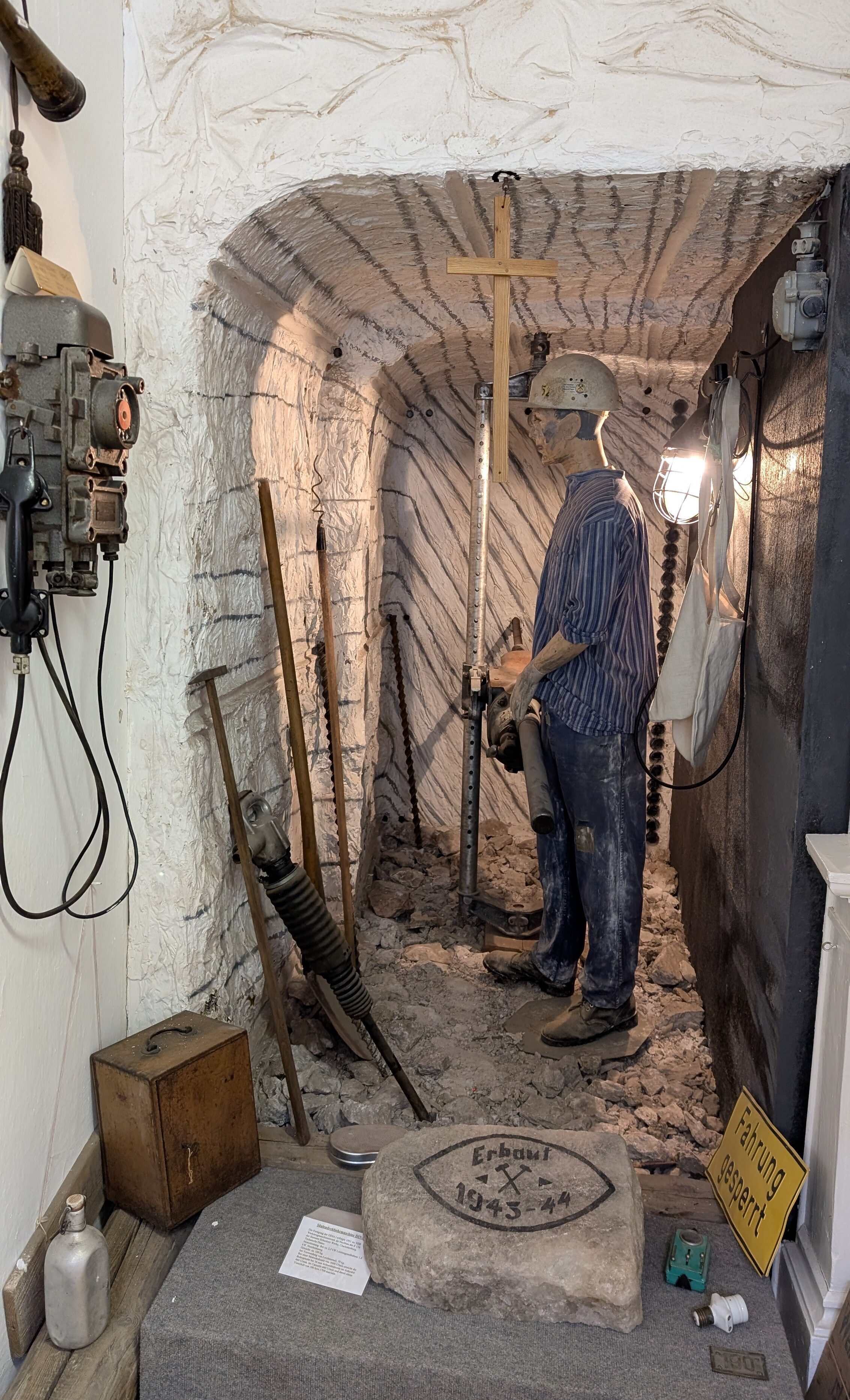 Die Rekonstruktion eines Salzschachtes mit einem Arbeiter und seinen benötigten Geräten. Der Arbeiter des Salzbergwerkes trägt einen Helm und dunkelblaue Arbeitskleidung. An der Wand hängt ein großes Telefon und daneben sind Arbeitsgeräte wie ein Bohrer und verschiedene Hacken aufgestellt.