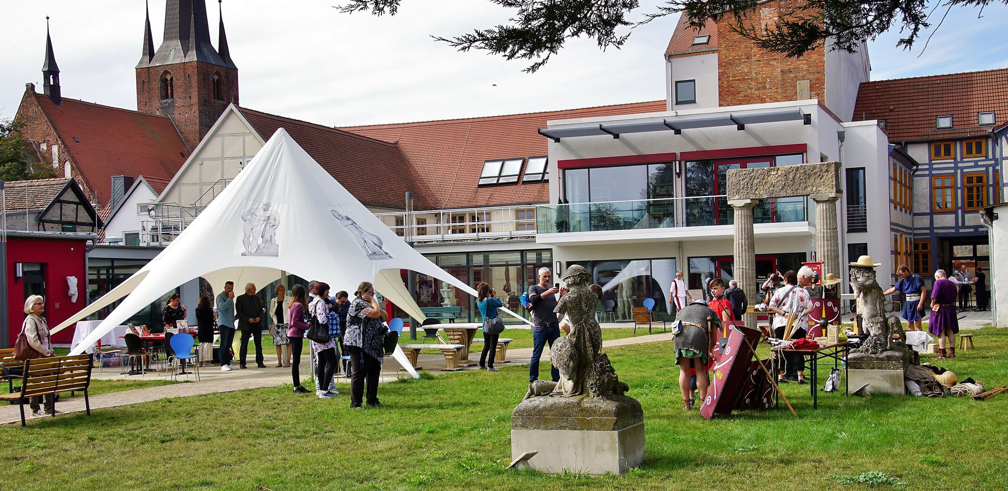 Das Winckelmann Museum in Stendal mit Winckelmann Park. Auf dem Rasen des Parks wird gerade ein Fest gefeiert. Zwischen den antik anmutenden Statuen im Park werde Kinder als Ritter mit Schilden verkleidet. Unter einem weißen Zelt sind erwachsene Menschen versammelt. Im Hintergrund befindet sich die teilweise verglaste und mit Fachwerk ausgeschmückte Fassade des Museum.