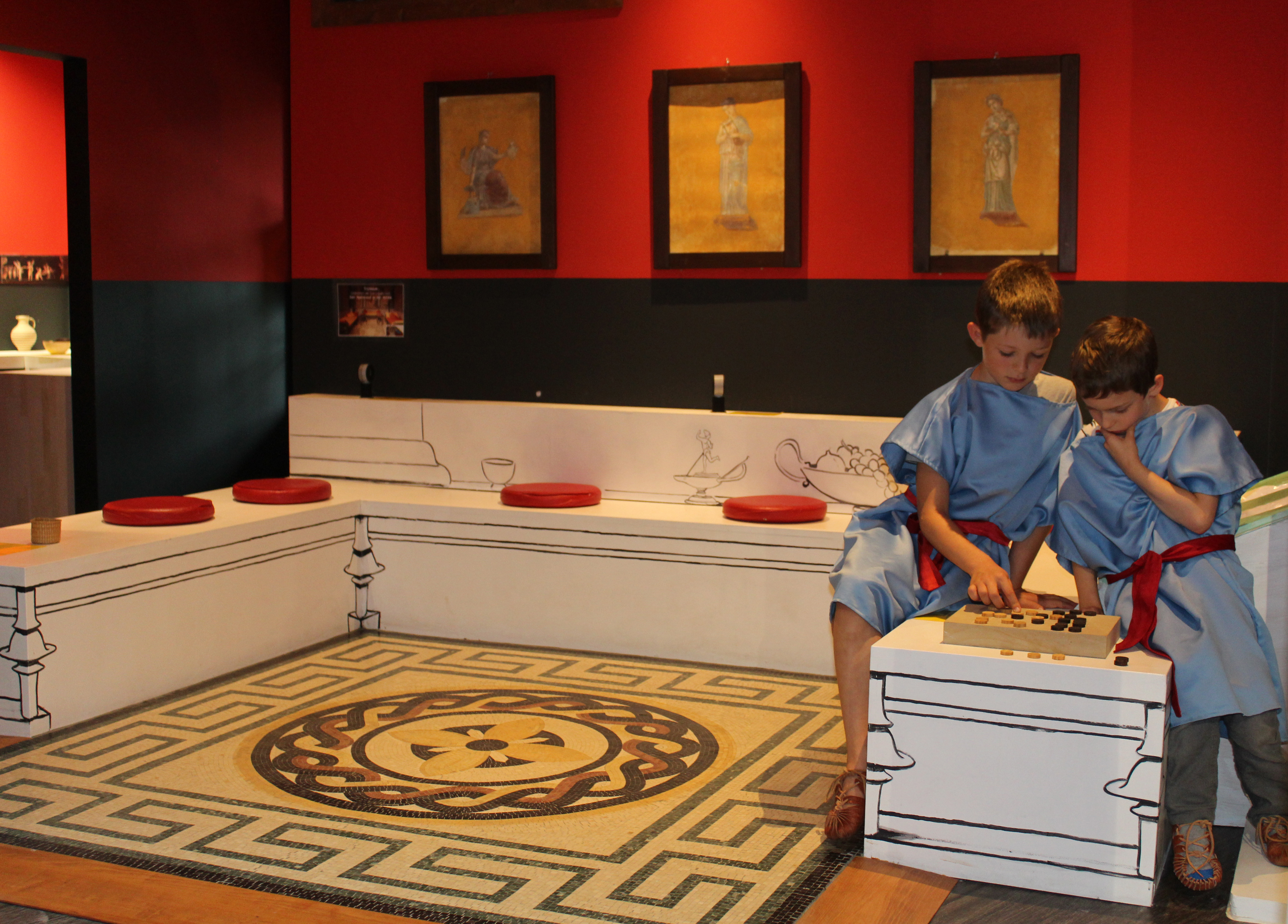 Familienmuseum im Winckelmann Museum. Zwei Kinder in einer blauen Tunika mit einem roten Gürtel sitzen auf einem weißen Sofa und spielen ein Brettspiel. Die Wände sind rot gestrichen und auf dem Boden liegt ein gelber Teppich, der an einen Mosaikfußboden erinnert.