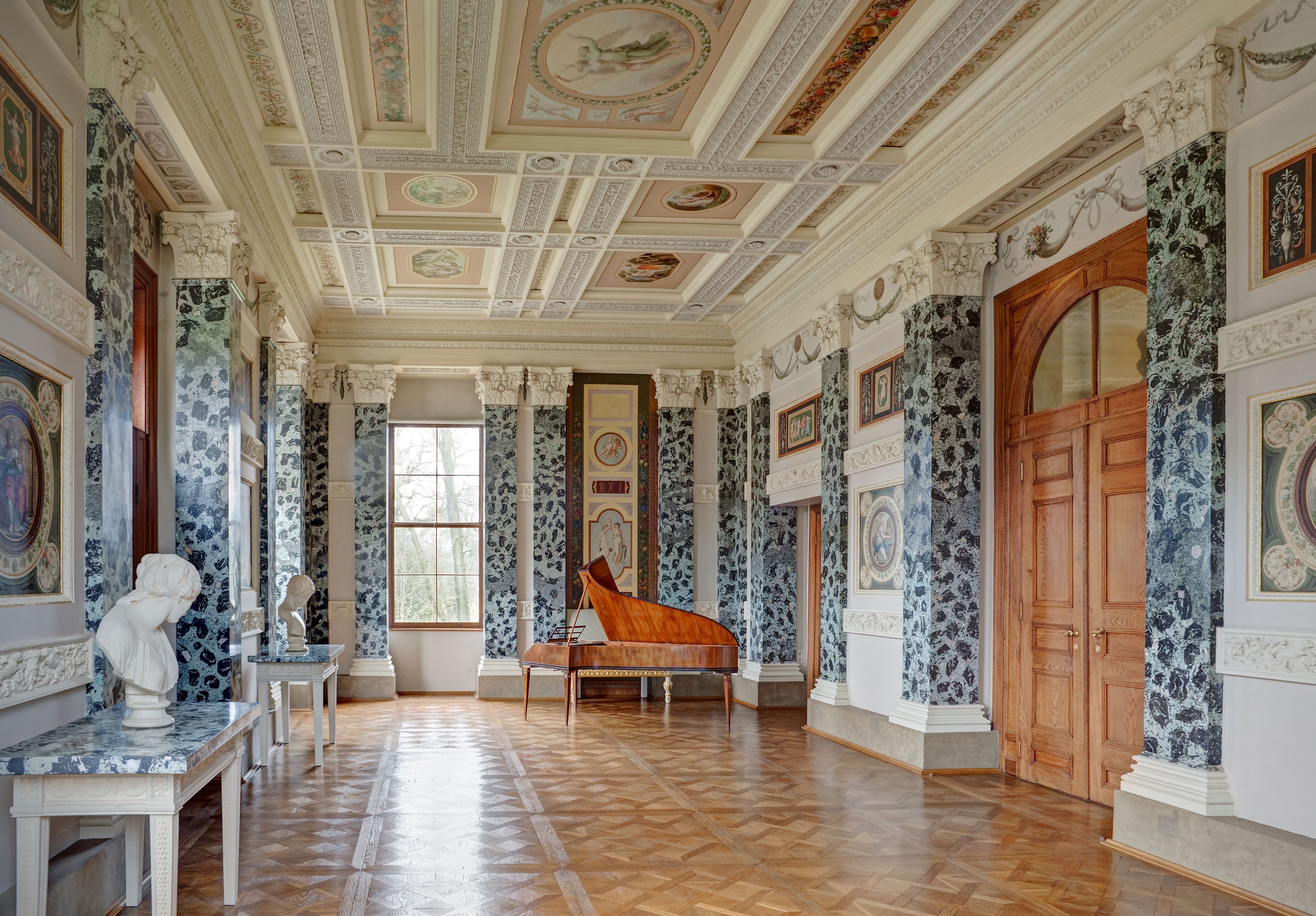 Der Festsaal im Schloss Luisium. Blau-schwarze Pilaster an den Wänden bestimmen den Raumeindruck. Die Decke und die Wänden sind mit weißem Stuck geschmückt. Am Ende des Saales steht ein großer Flügel.