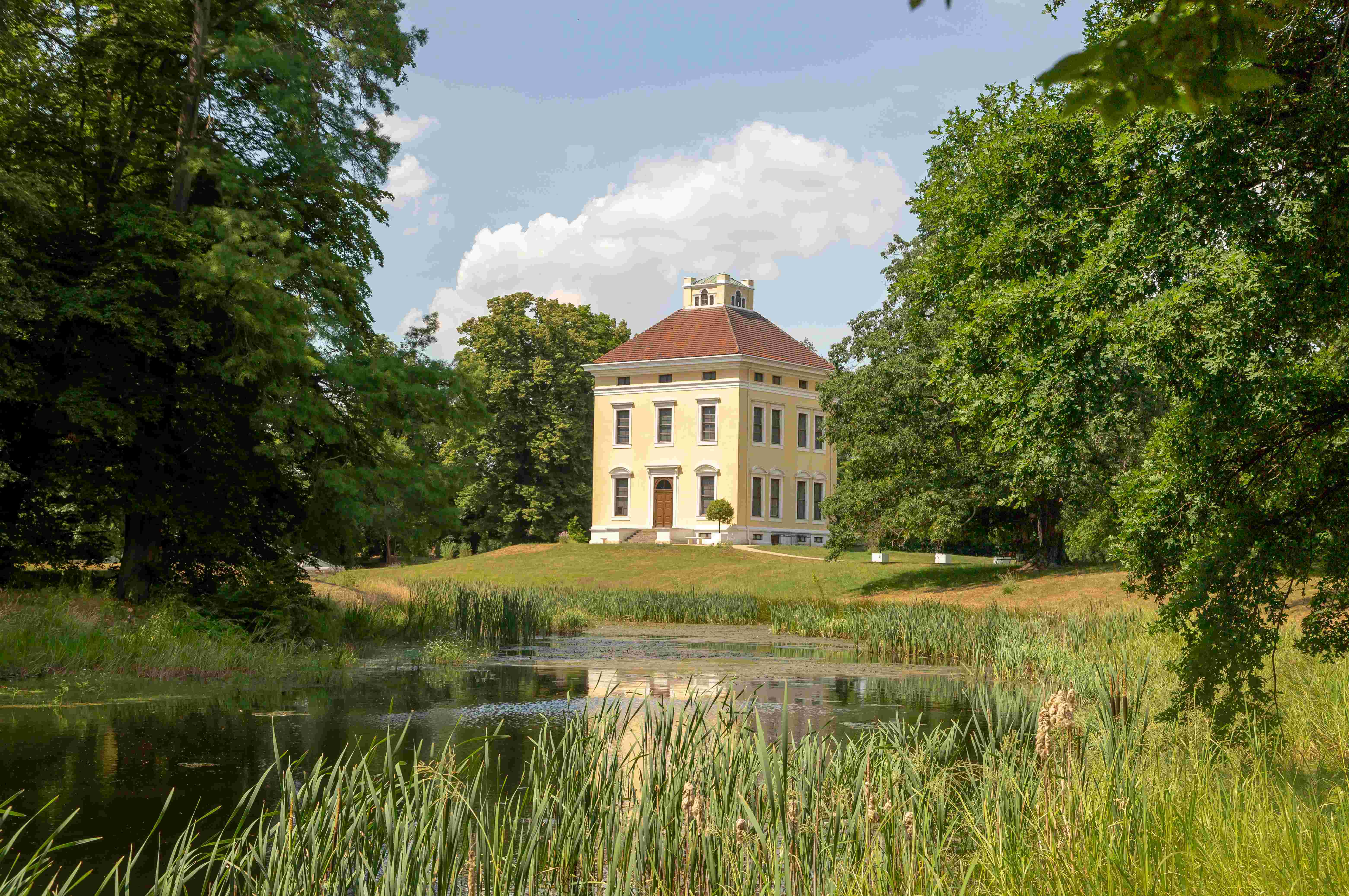 Das Schloss Luisium von Außen. In einer grünen Parkanlage mit einem See steht ein kleines Fürstenhaus mit gelber Fassade und einem roten Ziegeldach.